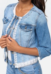 Chaqueta de mezclilla azul claro con escote redondo, borde de encaje floral y cierre frontal de botones, con dos bolsillos en el pecho.
