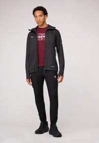 Zwarte softshelljas met een capuchon, ritszakken en een bordeauxrood graphic t-shirt eronder. Zwarte tapered broek en wandelschoenen.