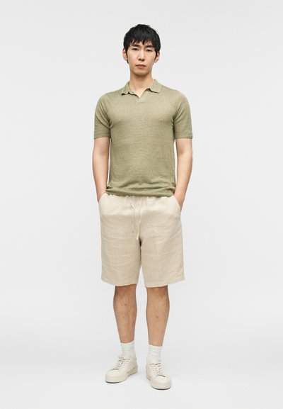 Πράσινο κοντομάνικο πόλο πουκάμισο συνδυασμένο με μπεζ λινό σορτς. Υπόδηση: ανοιχτόχρωμα αθλητικά παπούτσια. Το μοντέλο στέκεται όρθιο με τα χέρια στις τσέπες.