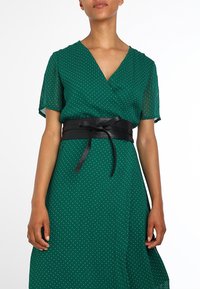 Green polka dot dress with a wrap design and short sleeves, accented by a black leather belt with ties. Smooth texture and flowy fabric.