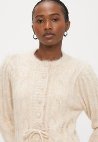 Κρεμ ζακέτα με πλέξη καλτσοδέτας, στρογγυλή λαιμόκοψη, κεντρικό κούμπωμα με κουμπιά και κορδόνι στη μέση. Μαλακό, υφασμάτινο με υφή.