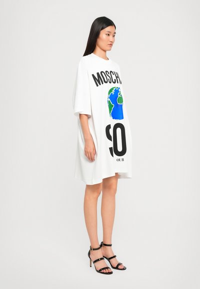 Weißes, oversized T-Shirt-Kleid mit "MOSCHINO"-Schriftzug und Globe-Grafik in Blau und Grün. Kombiniert mit schwarzen High-Heels.