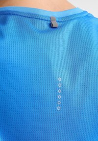 Blå kortärmad skjorta med texturerad tyg, som har en ögla vid halsen och fem små, runda detaljer längs ryggen.