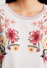 Blusa de base blanca con estampado floral, que presenta flores rojas, rosas y amarillas. Cuello texturizado con acentos acanalados.