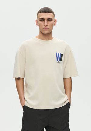 Młody mężczyzna o krótkich włosach, ubrany w beżową, oversize'ową koszulkę z niebiesko-czarnym napisem "WW" oraz czarne bermudy z kieszeniami, z rękami w kieszeniach.