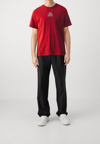 Röd bomullströja med Lacoste-logotyp på bröstet, i kombination med löst sittande svarta byxor och vita sneakers. Slät textur.