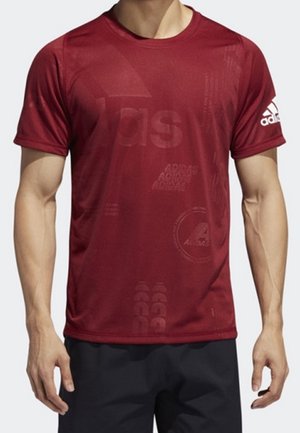 Homme portant un t-shirt rouge ajusté Adidas avec des motifs subtils du logo et un logo blanc Adidas sur la manche gauche, associé à un short noir.