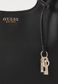 Sac à main en cuir noir avec une finition texturée, arborant un logo doré et des accents de porte-clés décoratifs. Haut arrondi et poignées robustes.