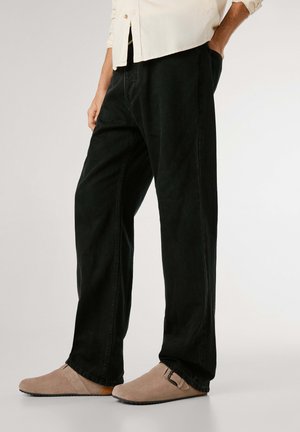 Pantalons en denim noir à coupe décontractée, avec des coutures minimales et des poches latérales. Associés à des chaussures à enfiler en suede beige avec une boucle.