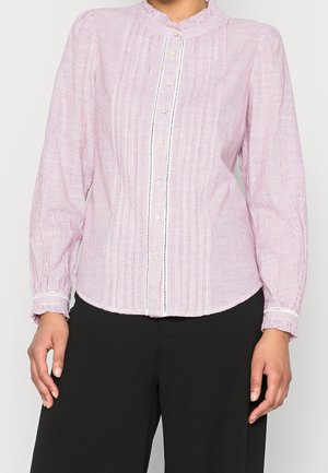 Femme portant un chemisier à manches longues à rayures rose clair et blanches avec un col à volants et des boutons en perles, associé à un pantalon noir.