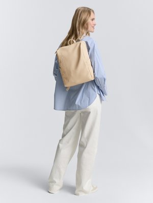 Mochila de lona beige con logo, forma rectangular, correas acolchadas y asa superior. Combinada con una camisa a rayas azul y pantalones blancos.