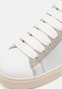 Gros plan sur la pointe d'une chaussure de sport blanche avec des lacets blancs épais, un panneau d'œillets en daim gris clair et une semelle extérieure texturée beige.