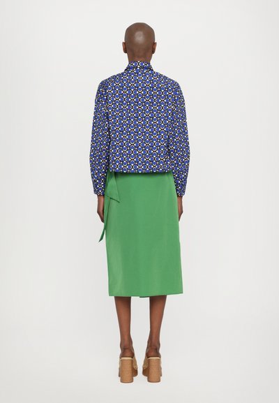 Cropped blue patterned jacket with abstract designs paired with a solid green skirt, featuring a tied accent at the waist. Brown platform heels.