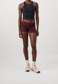 Débardeur de sport noir avec des accents roses, short bordeaux avec taille élastique « NIKE », chaussettes blanches et baskets beige. Tissu lisse et ajusté.