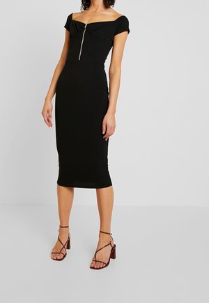 Femme portant une robe midi noire épaules dénudées avec fermeture éclair devant et des sandales marron à talons hauts à brides, debout devant un fond uni.
