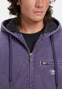 Purple hoodie with black quilted lining, featuring a zippered front, drawstrings, and a chest pocket with a decorative logo on fabric.