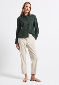 Donkergroen blouse met bloemmotieven, golvende zoom, knoopsluiting; gecombineerd met lichtbeige broek en blootvoetse sandalen.