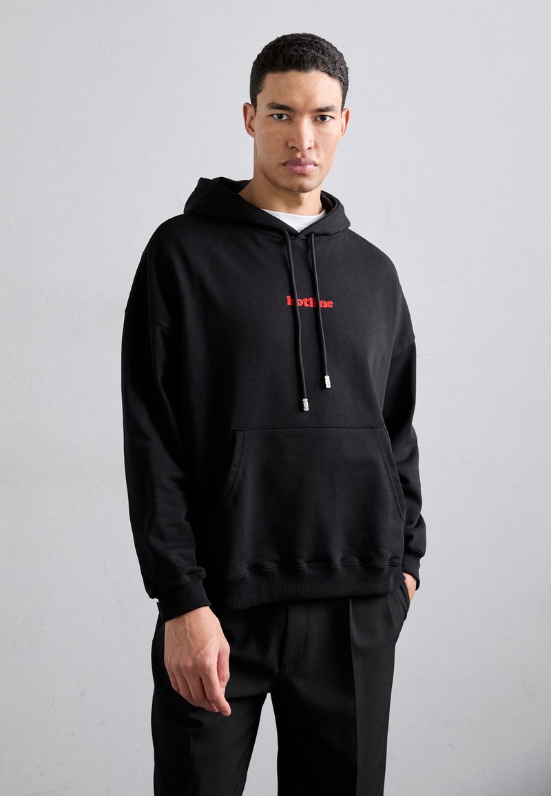 Črna kapuca, izdelana iz mehkega materiala, z sprednjim žepom in rdečim napisom "hotline" na prsih. Detajli z vrvico pri kapuci.