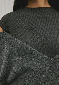Dunkelgrauer Rippstrickpullover mit hohem Kragen, unter einem weichen, fusseligen Cardigan in Anthrazit getragen, der verschiedene Texturen und überlappende Designs präsentiert.
