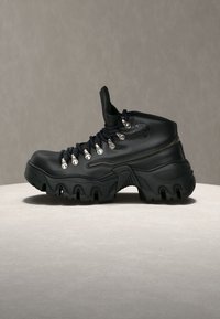 Botte de randonnée montante en cuir noir avec semelle épaisse et robuste, lacets noirs, œillets argentés, logo embossé à la cheville, présentée sur une surface neutre.