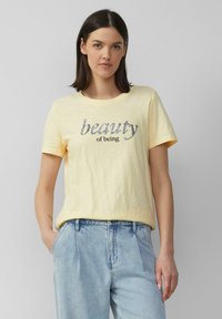 T-shirt en coton jaune clair avec une coupe décontractée, un col rond et un graphisme "la beauté d'être" en argent et noir. Porté avec un jean bleu clair.