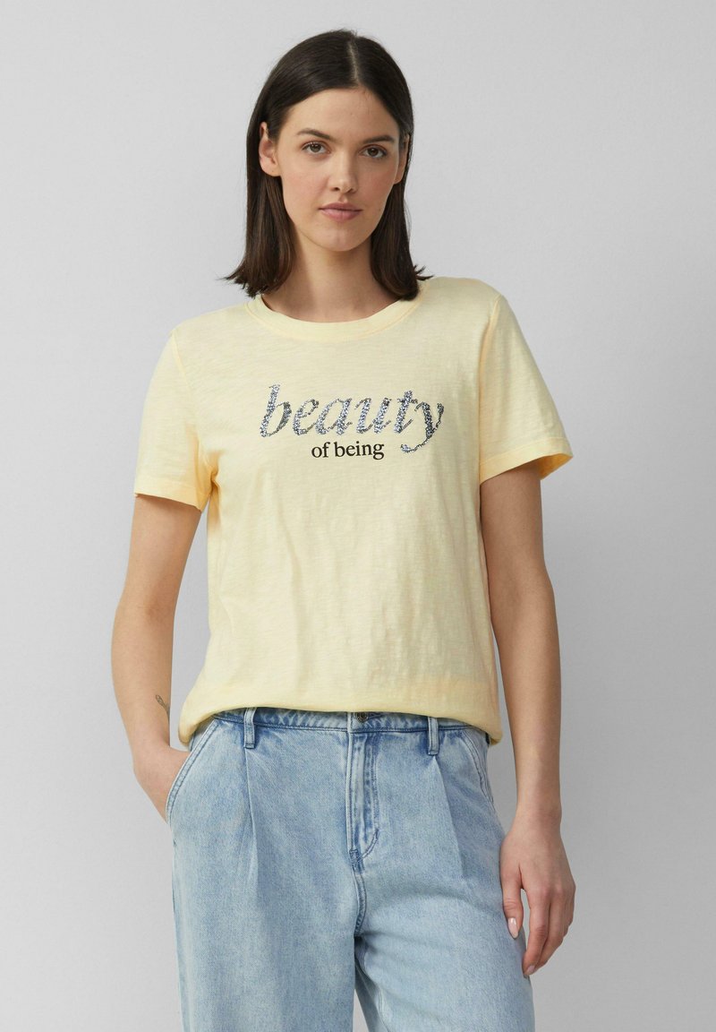 T-shirt en coton jaune clair avec une coupe décontractée, un col rond et un graphisme "la beauté d'être" en argent et noir. Porté avec un jean bleu clair.