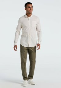 Lichtbeige overhemd met knopen en een zak; gecombineerd met olijfgroene cargobroek. Model staat en draagt witte sneakers.