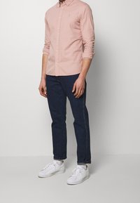 Camisa de botones de color rosa claro con cuello, combinada con jeans azul oscuro y zapatillas blancas. Corte casual con mangas remangadas. Detalle sutil de logo.