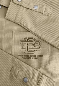 Beige, gestructureerde stof met een diagonale ribpatroon. Beschikt over een borstzak met een gebosst logo en zilveren drukknoopjes.