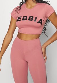 Lyserød cropped sports top med sort skrivning "EBBIA," parret med matchende højtaljede leggings; glat, strækbart stof, tætsiddende design.