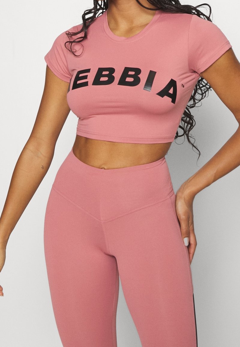 Lyserød cropped sports top med sort skrivning "EBBIA," parret med matchende højtaljede leggings; glat, strækbart stof, tætsiddende design.