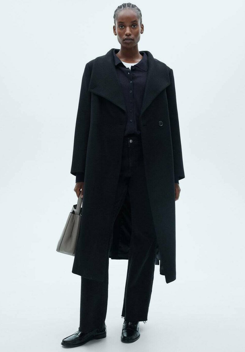 Manteau en laine noir avec grands revers, fermeture à boutons, porté sur une chemise sombre et un jean noir. Un sac à main gris et des chaussures noires complètent le look.