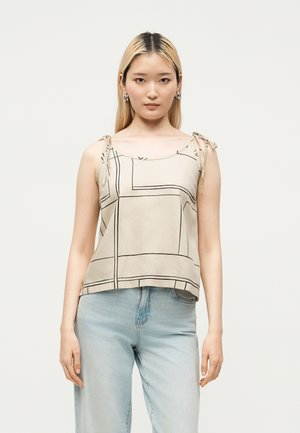 Femme aux longs cheveux blonds porte un haut beige sans manches avec de fines bretelles et des lignes géométriques noires, associé à un jean bleu clair.