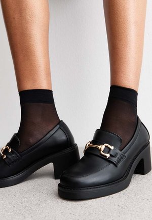Svarta läderplatform-loafers med rundad tå, med detalj i guldtonad spänne och tjocka, texturerade klackar. Svarta genomskinliga strumpor bärs.