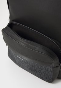 Zaino nero con parte superiore in pelle liscia e tasca con zip, e un compartimento inferiore stampato e testurizzato che mostra un motivo logo.