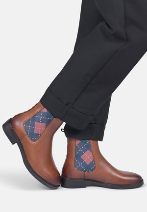 Brown leather Chelsea boots with blue and pink argyle elastic side panels worn with black cuffed pants.