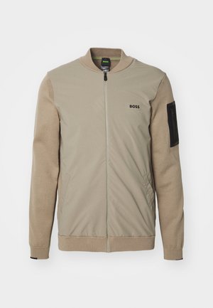 Beige jakke med ribbet krage og mansjetter. Har glidelåslukking, sidelommer, og en svart glidelåslomme på venstre erme.