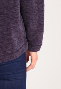 Pullover in tessuto porpora, con texture, orlo stondato e dettagli di cucitura visibili; la mano appoggiata al fianco contro un jeans scuro.