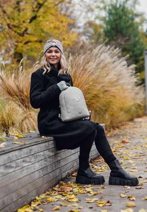 Grauer Rucksack mit sechseckigem Muster und V-Logo, kombiniert mit einem schwarzen Mantel, schwarzen Leggings und klobigen schwarzen Plateau-Stiefeln. Herbstliche Szenerie.