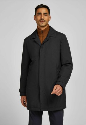 Schwarzer Mantel mit strukturiertem Design, breitem Kragen, Seitentaschen und durchgeknöpfter Front, getragen über einem braunen Rollkragenpullover.