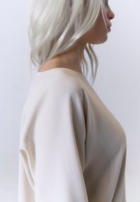 Beige, strukturiertes Bluse mit lockerer Passform und weitem Ausschnitt. Der Stoff fällt sanft und zeigt leichte Bewegungen sowie eine glatte Oberfläche.