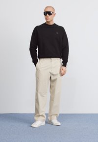 Svart hettegenser med en liten logo, beige løstsittende bukser, og hvite sneakers. Personen har på seg store svarte solbriller.