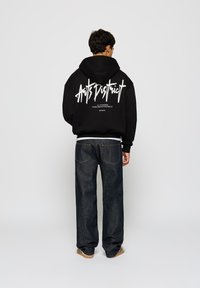 Svart hettegenser med hvit grafisk tekst på ryggen, sammen med mørk denim jeans og brune joggesko. Uformell passform, minimal detaljering.