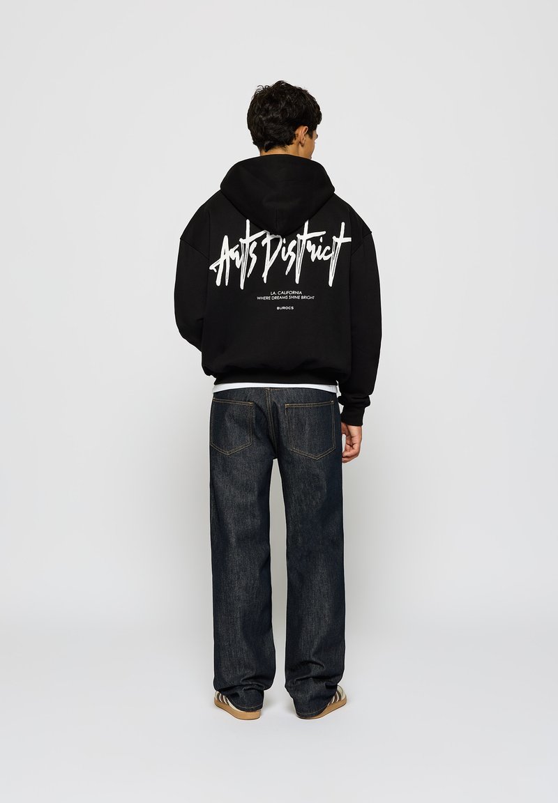 Svart hettegenser med hvit grafisk tekst på ryggen, sammen med mørk denim jeans og brune joggesko. Uformell passform, minimal detaljering.