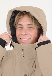 Beige vattentålig jacka med huva, med svarta detaljer, frontdragkedja och justerbara ärmslut. Slät textur och varm foder synligt.