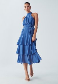Plava čipkasta haljina s halter vratom, slojevitim volanima na donjem rubu i odgovarajućim remenom vezanim oko struka, uz bež sandale s trakicama.