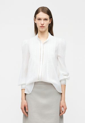 Woman wearing a white long-sleeve blouse with lace details and a light gray skirt, standing against a plain light background.