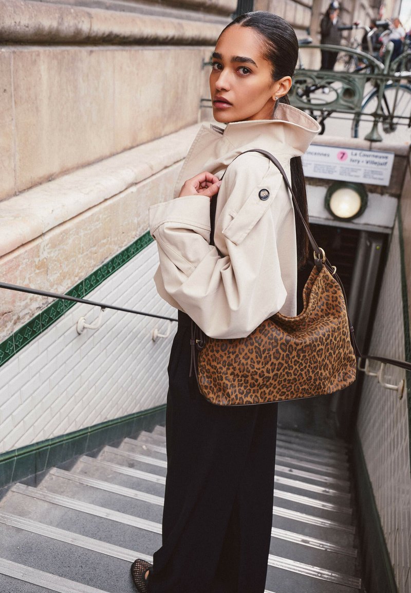 Manteau beige avec un col montant, pantalon noir à jambes larges, et un sac à main hobo léopard marron et noir, avec des marches en arrière-plan.