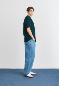 Temnozeleni kratki majica s kratkimi rokavi, v kombinaciji z ohlapnimi svetlo modrimi denim hlačami; model stoji bočno, z rokami v žepih, na nogah ima bele superge.