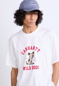 White cotton t-shirt features a cartoon dog with red "CARHARTT WILD DOGS" text. Gray cap with logo, short sleeves, relaxed fit.